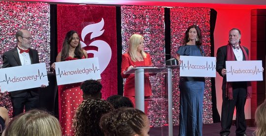 A blonde woman is in the middle of the image at a podium in front of different red backgrounds. There are two people on the left of her holding signs that say "discovery" and "knowledge", and two people on the right holding signs that say "access" and "advocacy".