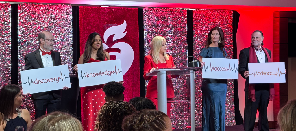 A blonde woman is in the middle of the image at a podium in front of different red backgrounds. There are two people on the left of her holding signs that say "discovery" and "knowledge", and two people on the right holding signs that say "access" and "advocacy".