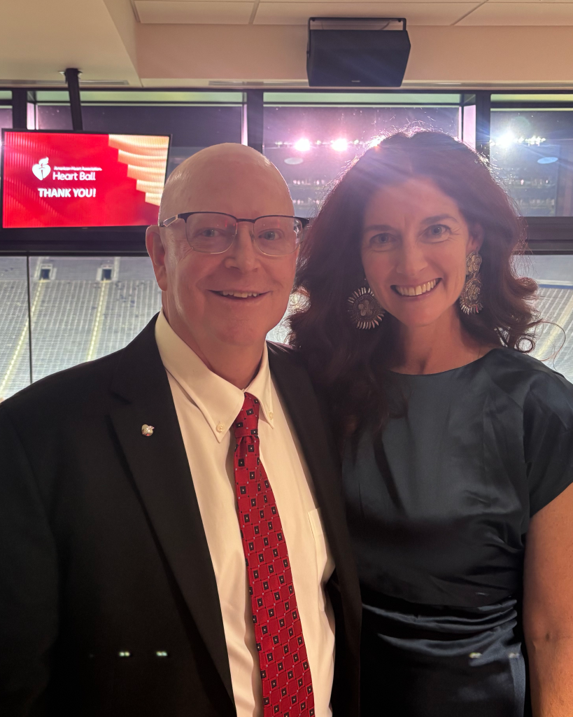 Bald man in suit standing next to a brown-haired woman in a blue dress. The AHA Heart Ball logo is on a screen behind them.